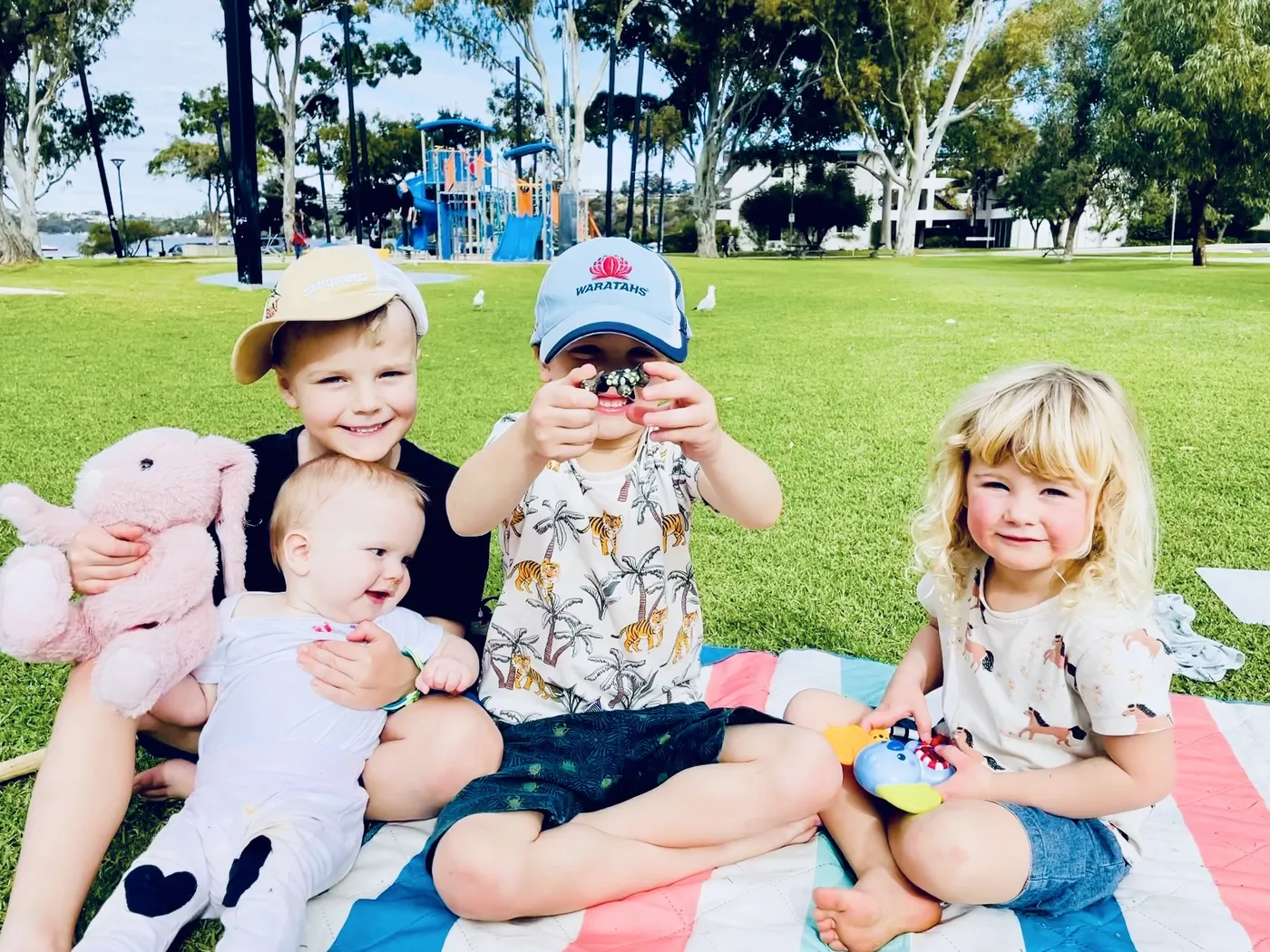 Four small children on a picnic rug in a park, laughing.